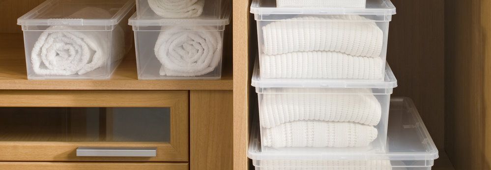 Ordentlich gefaltete Kleidung und Handtücher in durchsichtigen Plastikbehältern in einem Holzschrank.